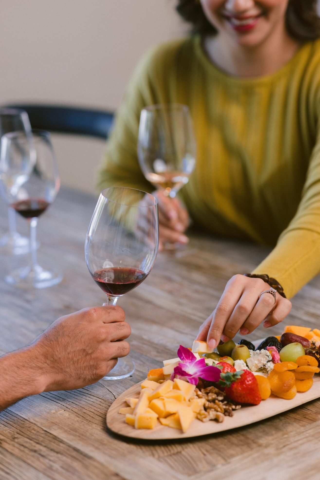 Two people sit at a wooden table with glasses of red wine, sharing a charcuterie board with cheese, grapes, nuts, and fruit. One person reaches for food while smiling. The mood is casual and friendly.