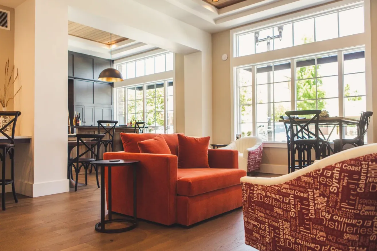 A bright, modern lounge area with a large orange armchair, patterned chairs, wooden floors, and several tables and chairs near large windows letting in natural light.