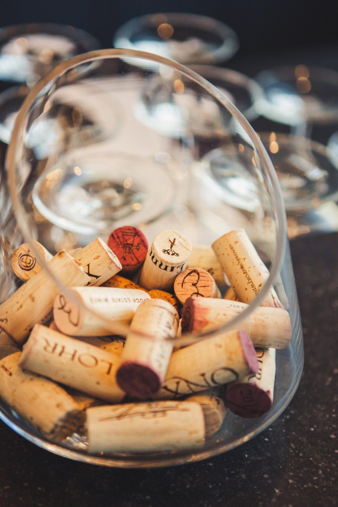 A glass container tipped on its side is filled with assorted wine corks, some with red wine stains. Empty wine glasses are blurred in the background on a dark surface.