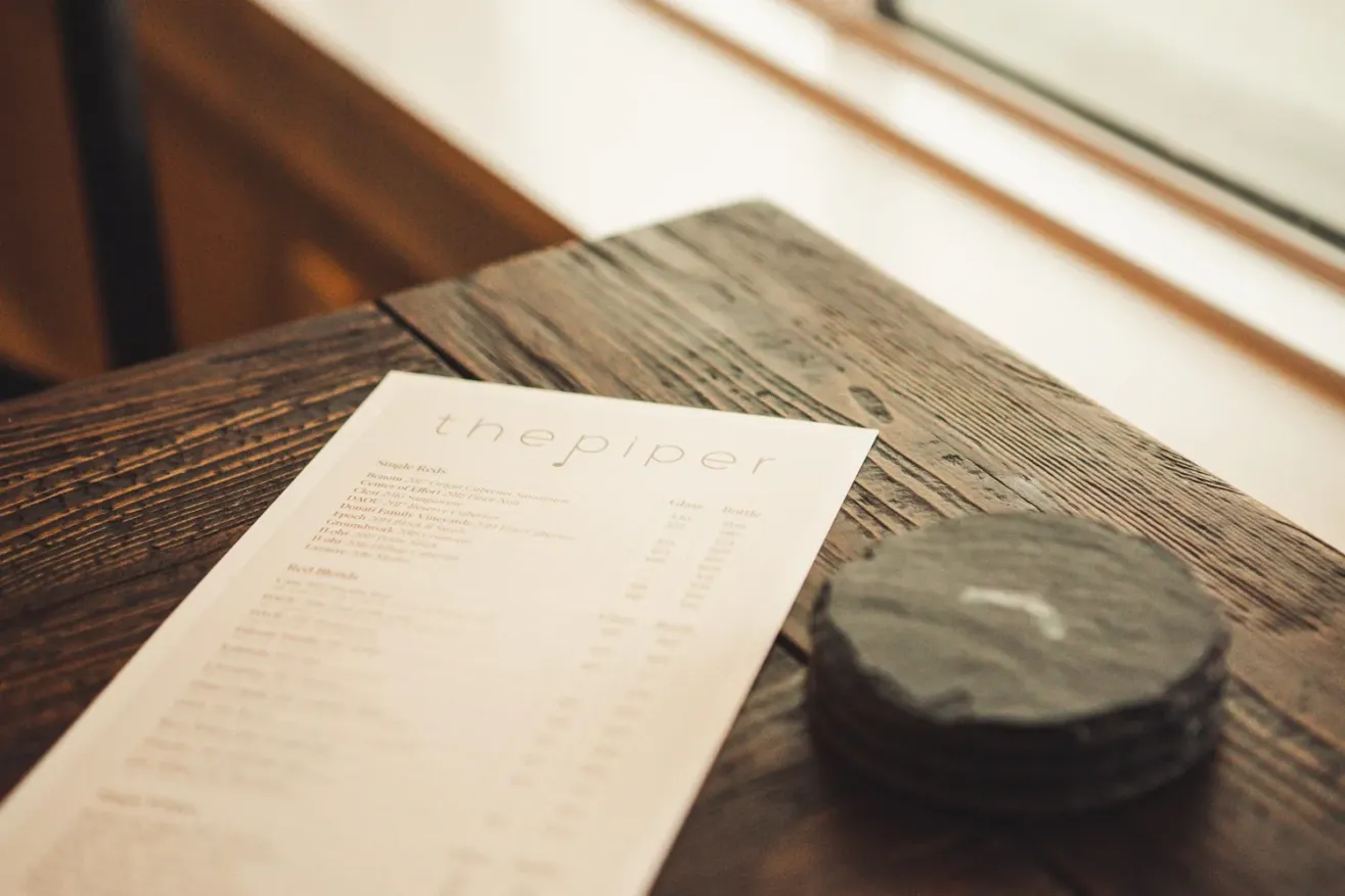 A restaurant menu titled the piper lies on a rustic wooden table next to a small stack of round, dark drink coasters by a window.