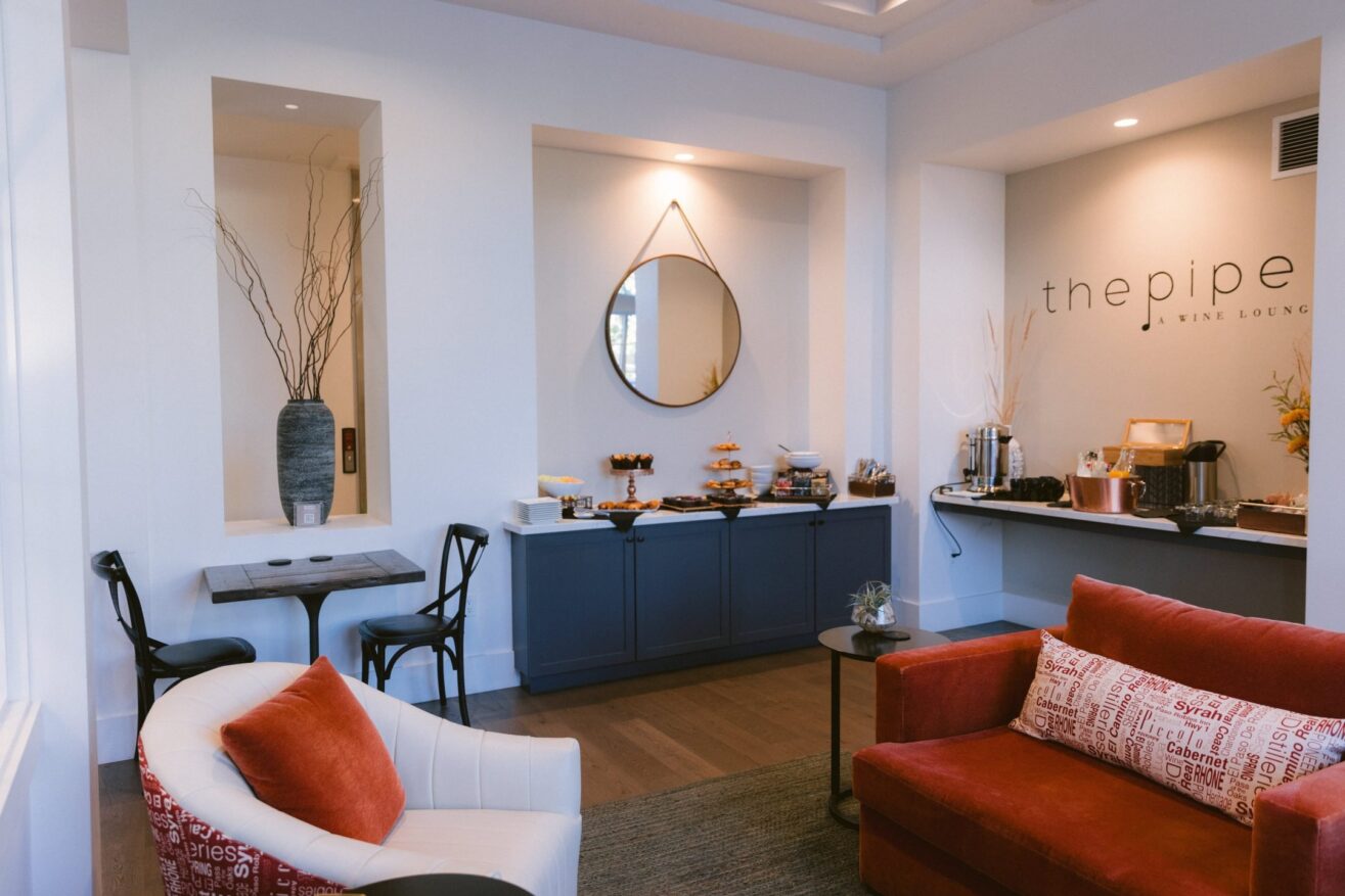 A modern wine lounge with a cozy seating area, red and white chairs, a display of snacks on blue cabinets, a round mirror, minimalist decor, and “the pipe WINE LOUNGE” written on the wall.