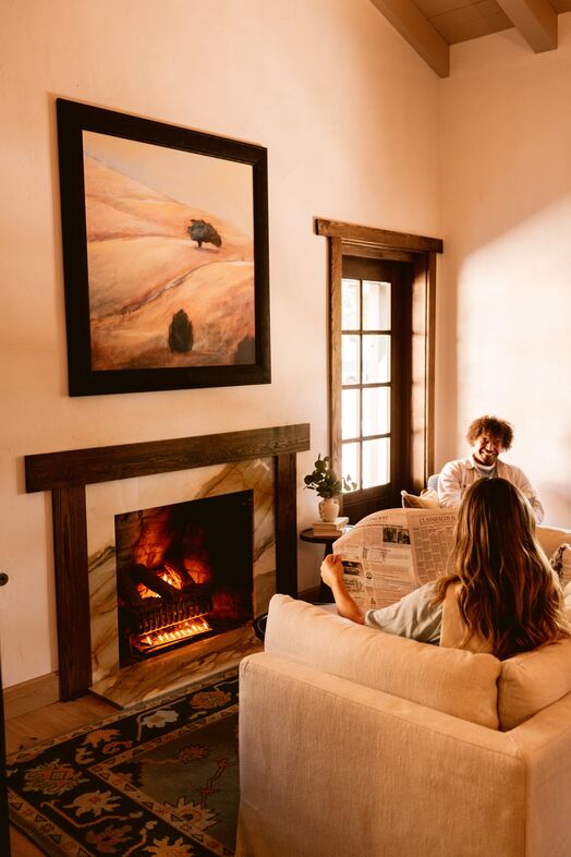 A cozy living room with a lit fireplace, a large landscape painting above it, and two people relaxing on sofas—one reading a newspaper and the other smiling. Warm sunlight streams through a window and glass door.