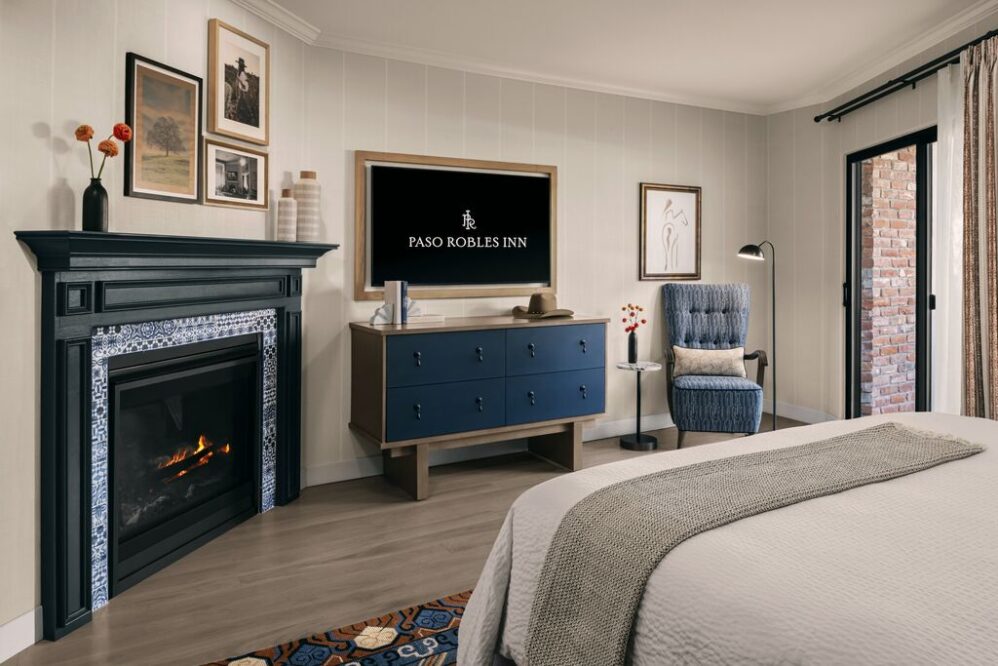 A cozy hotel room with a bed, fireplace, dresser with a TV displaying Paso Robles Inn, an armchair with a lamp, framed wall art, and a door leading to a brick patio. The decor is modern and inviting.