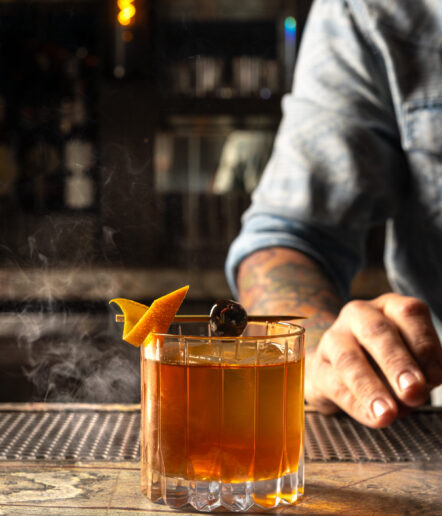 A person in a denim shirt holds a glass of amber cocktail on a bar, garnished with an orange twist and a cherry. Wisps of smoke rise from the drink. The background is dimly lit and out of focus.