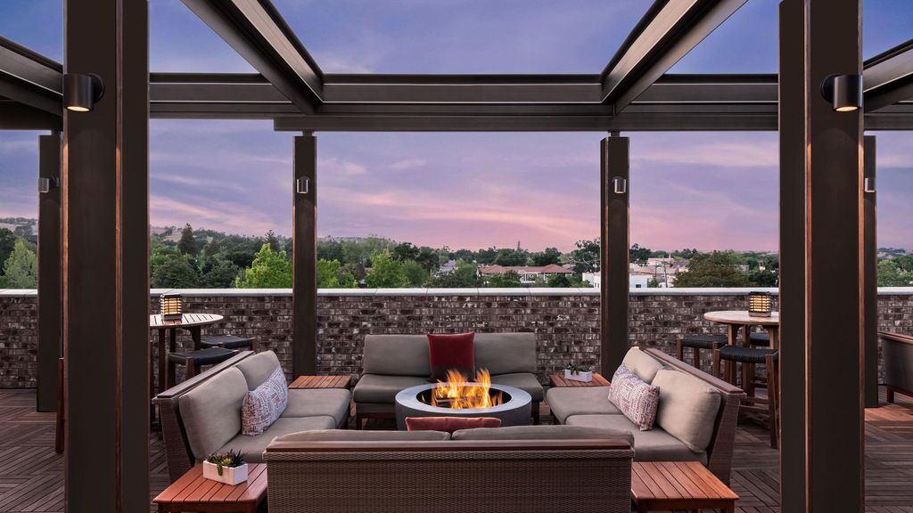 Outdoor rooftop lounge at Luxury Hotel Paso Robles features cushioned seating around a fire pit under a pergola. Tall tables and chairs invite you to enjoy sunset views over trees and houses beneath a purple and pink sky.