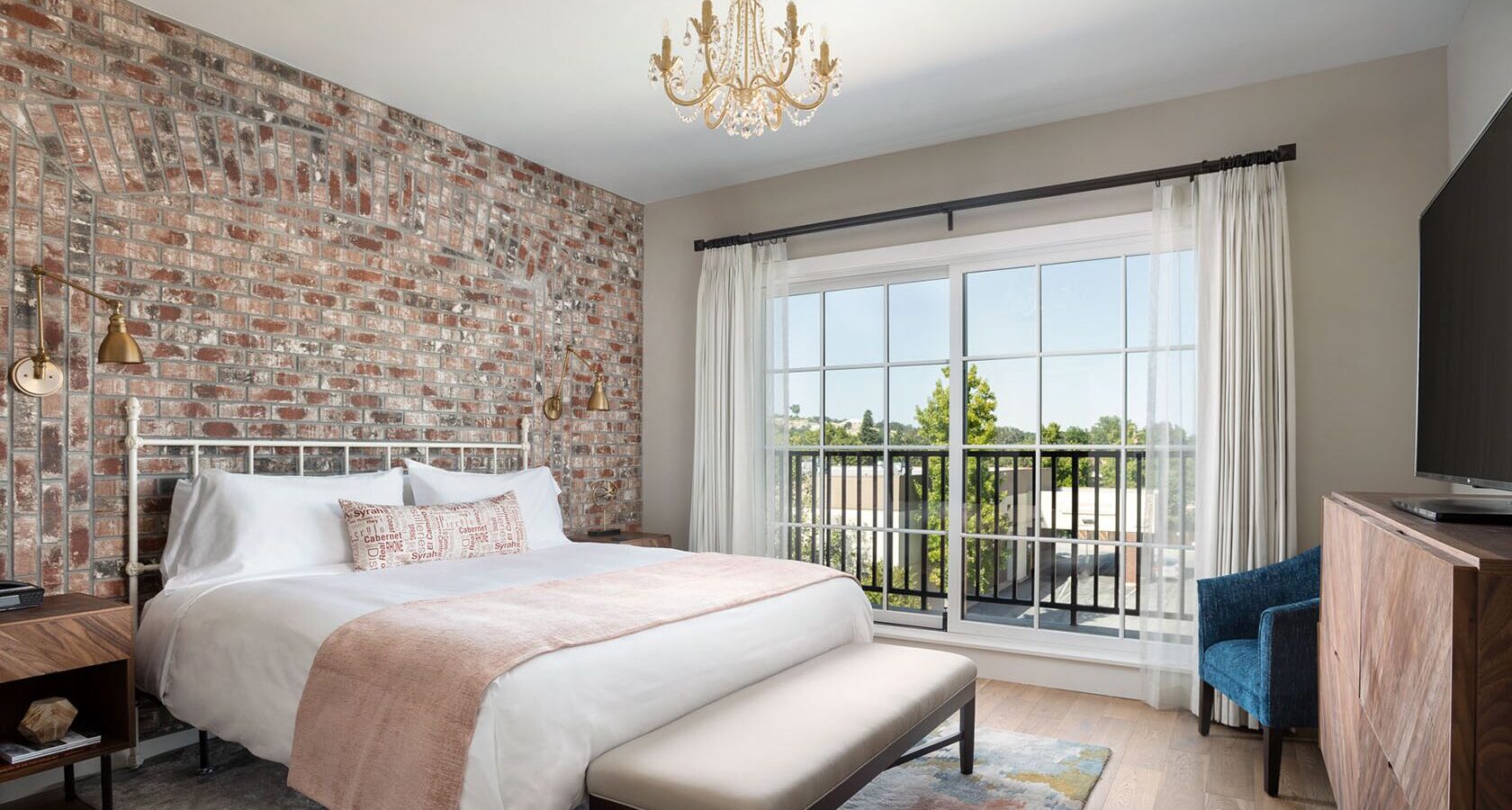 A modern bedroom at Luxury Hotel Paso Robles features a brick accent wall, white bed, beige bench, wooden furniture, blue armchair, chandelier, large window with white curtains, and a view of trees outside.