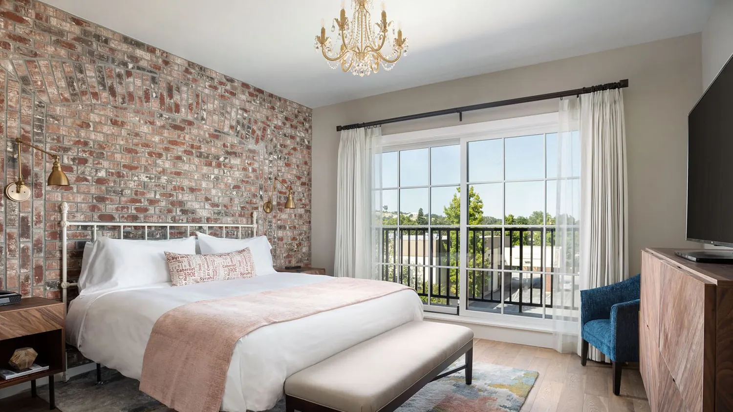 A modern bedroom at Luxury Hotel Paso Robles features a brick accent wall, white bed, beige bench, wooden furniture, blue armchair, chandelier, large window with white curtains, and a view of trees outside.