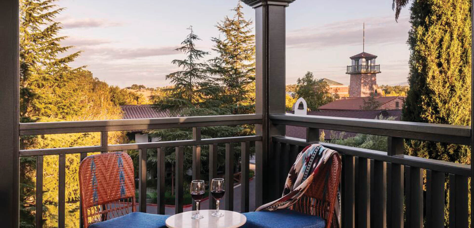 Two wicker chairs with blue cushions and a small table holding two glasses of red wine sit on a balcony overlooking trees and a distant tower at sunset. A patterned blanket drapes over one chair.