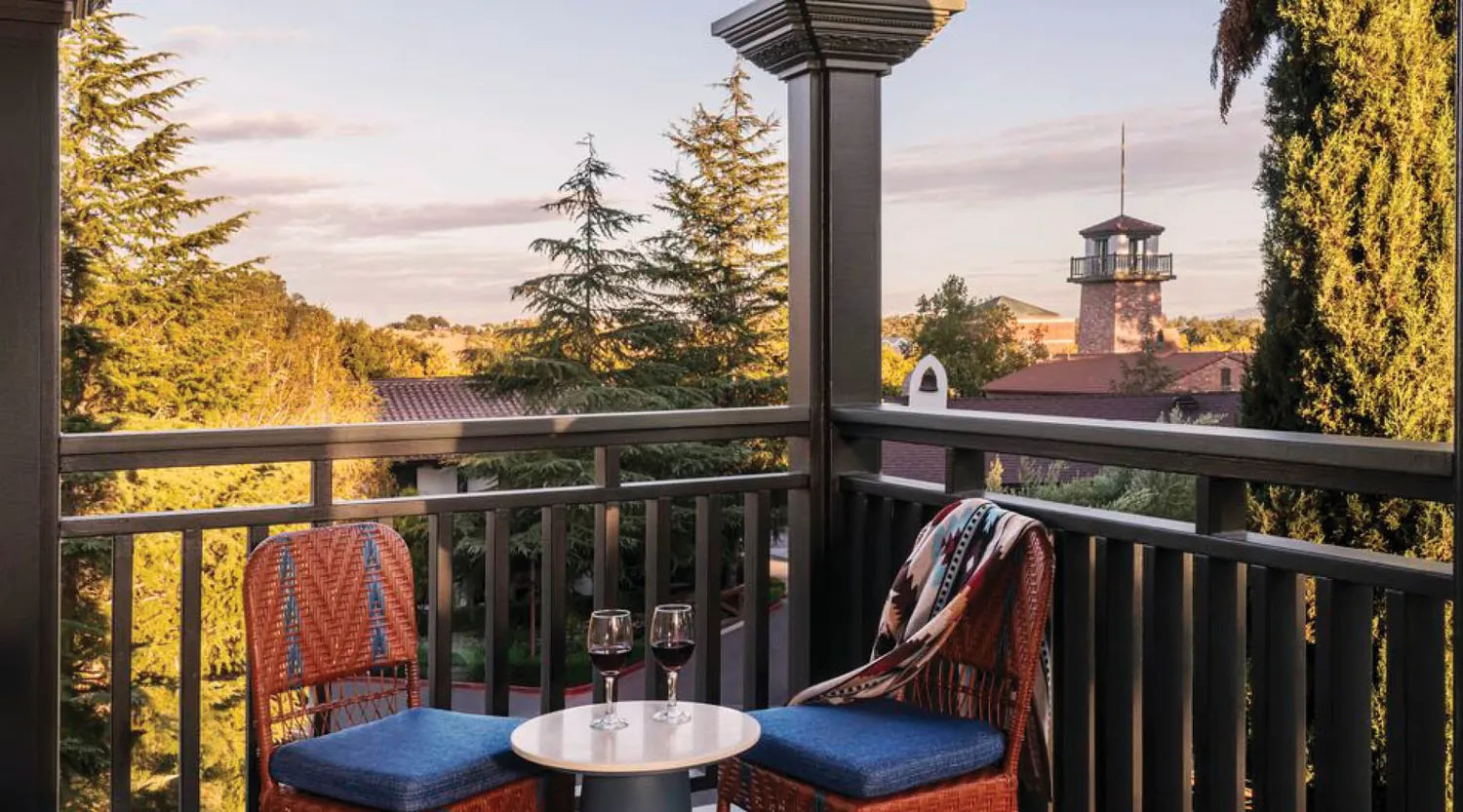 Two wicker chairs with blue cushions and a small table holding two glasses of red wine sit on a balcony overlooking trees and a distant tower at sunset. A patterned blanket drapes over one chair.