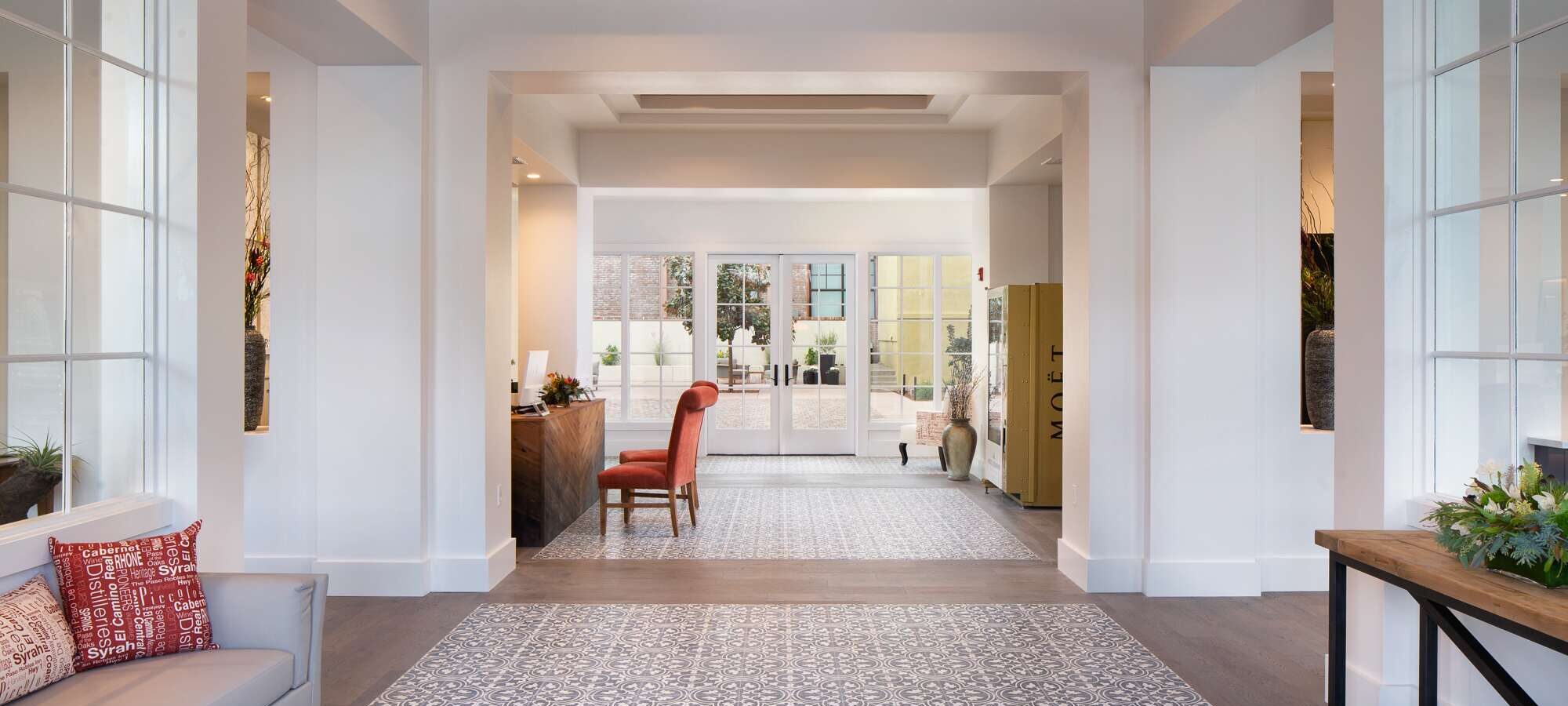 Bright, modern lobby at The Piccolo at Paso Robles Inn with patterned tile floor, a red accent chair at a wooden desk, large windows flooding the space with natural light, contemporary décor, lush plants and a cushioned bench adorned with red pillows.