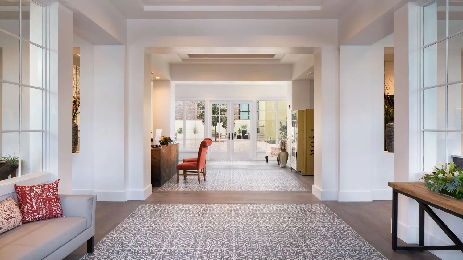 Bright, modern lobby at The Piccolo at Paso Robles Inn with patterned tile floor, a red accent chair at a wooden desk, large windows flooding the space with natural light, contemporary décor, lush plants and a cushioned bench adorned with red pillows.