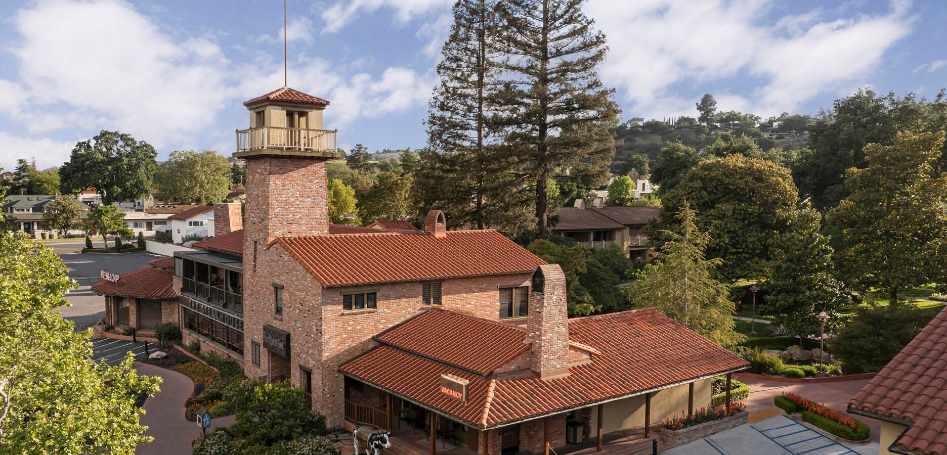 Aerial view of a two-story brick building with a tall tower, red tile roof, chimneys, and surrounded by lush green trees and landscaped pathways under a partly cloudy sky.