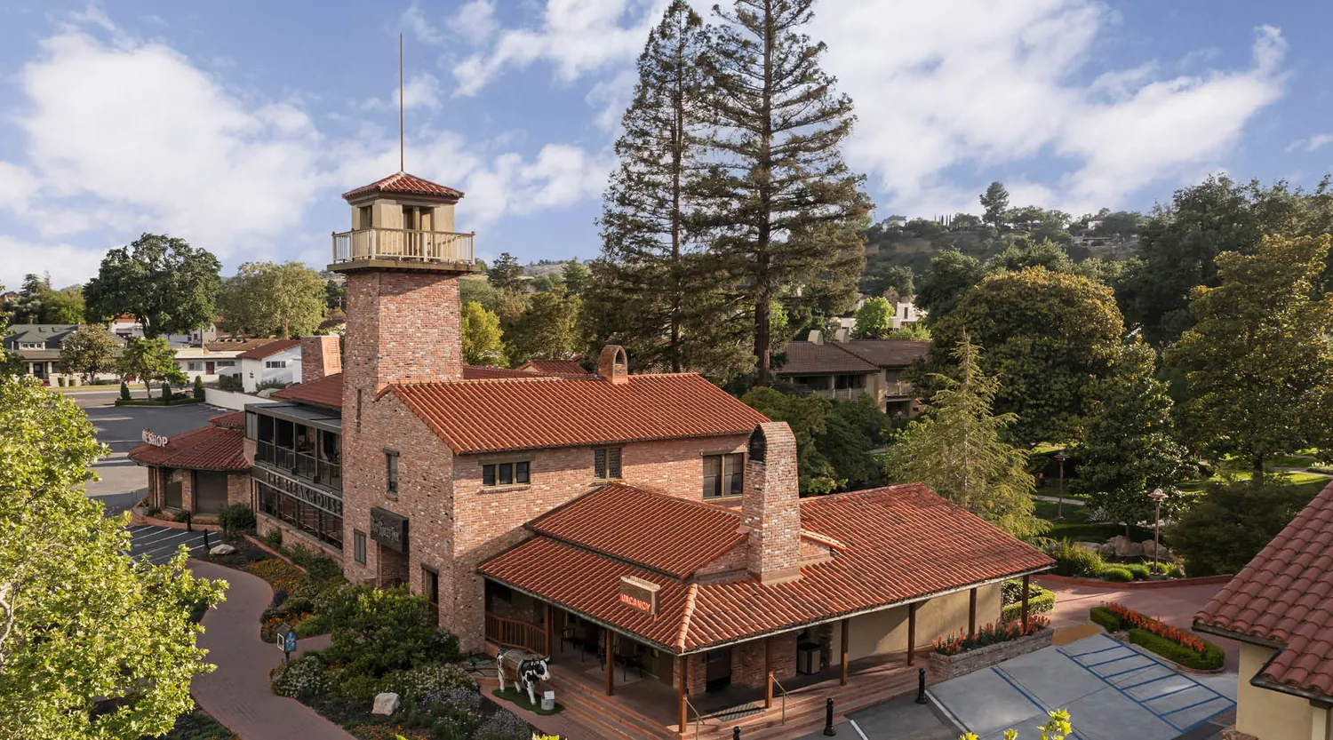 Aerial view of a two-story brick building with a tall tower, red tile roof, chimneys, and surrounded by lush green trees and landscaped pathways under a partly cloudy sky.