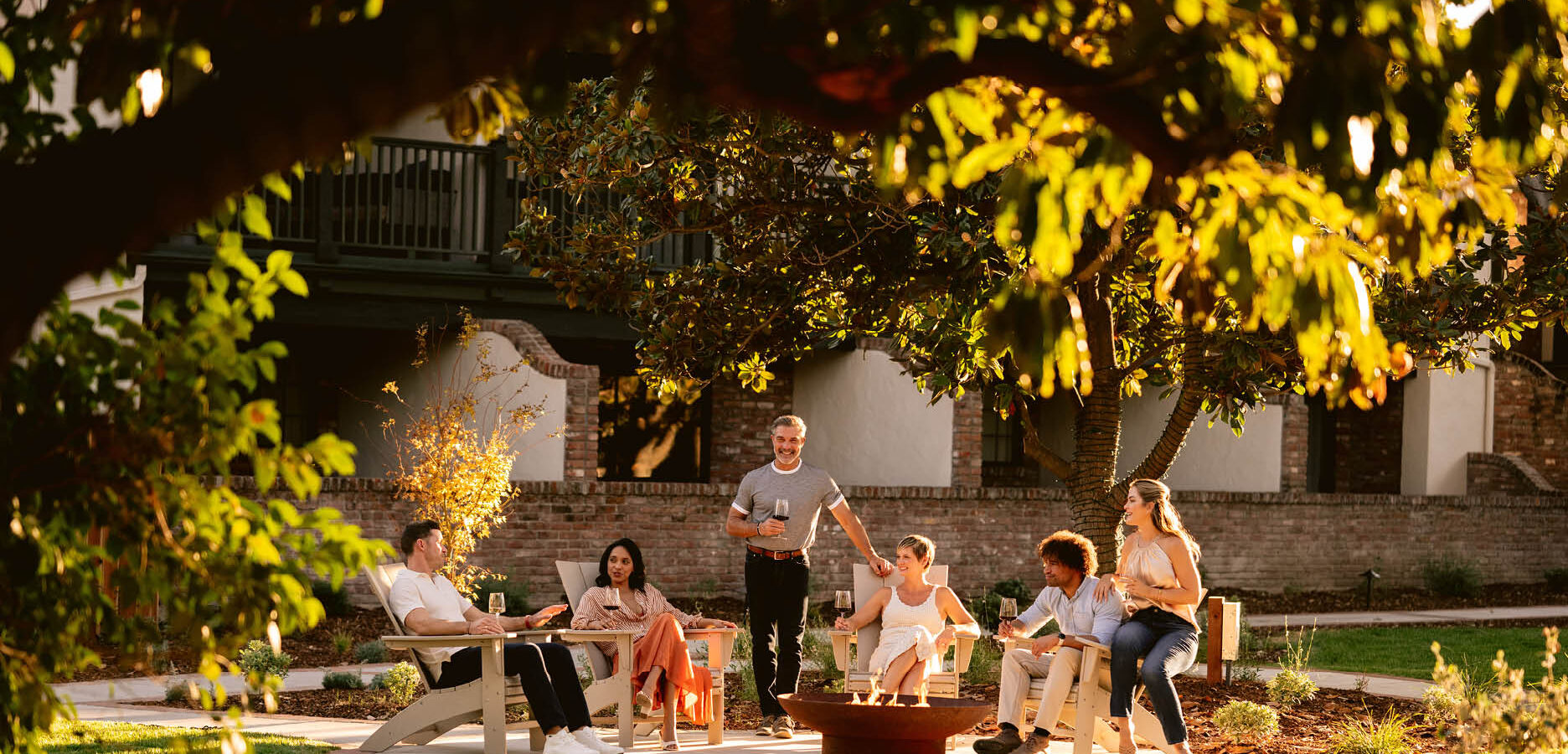 A group of six people sit and stand around a fire pit in a sunny outdoor garden, talking and relaxing on chairs, surrounded by trees and greenery with a building in the background.