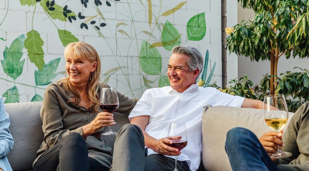 Two people sit on an outdoor couch, smiling and holding glasses of wine. Green plants and a mural of leaves decorate the background, creating a relaxed, cheerful atmosphere.