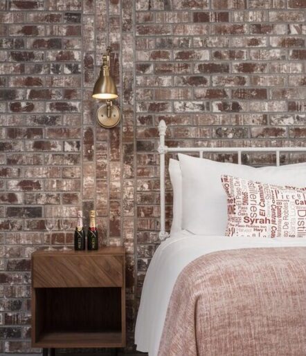 A cozy bedroom with an exposed brick wall, a white metal bed frame, white bedding, a patterned pillow, a wooden nightstand with wine bottles and glasses, and a wall-mounted brass light fixture.