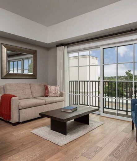 Modern living room with a beige sofa, red throw blanket, and a large mirror on the wall. A black coffee table sits on a rug near sliding glass doors that open to a balcony with a view of trees and buildings.