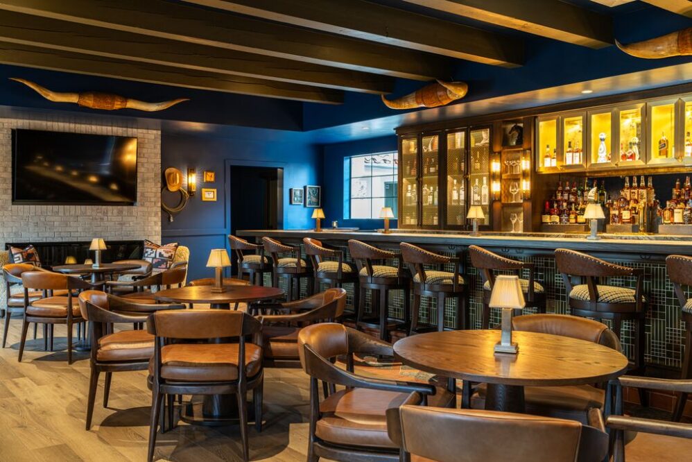 A stylish bar interior with wooden tables and chairs, a long bar counter lined with stools, illuminated shelves with bottles, a large TV, and decorative bull horns on dark blue walls. Warm lighting creates a cozy atmosphere.