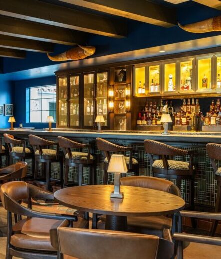 A stylish bar with dark wooden chairs and tables, a marble countertop, warm lighting, and shelves stocked with bottles behind the bar. Small lamps sit on the bar and tables, creating a cozy, inviting atmosphere.