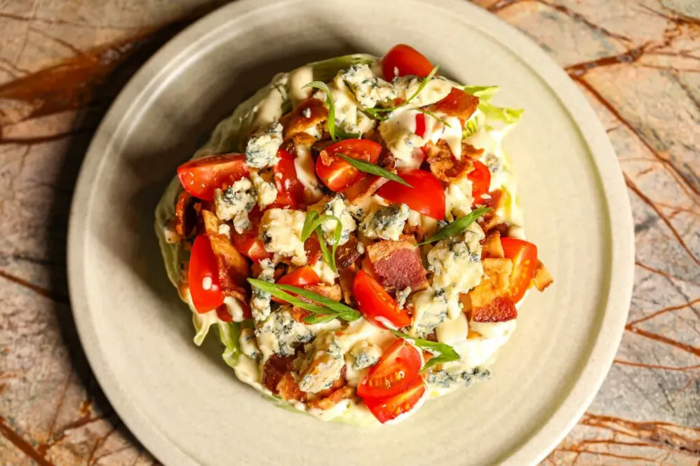 A wedge salad topped with cherry tomatoes, blue cheese crumbles, crispy bacon, chopped green onions, and creamy dressing, served on a beige plate over a marbled surface.