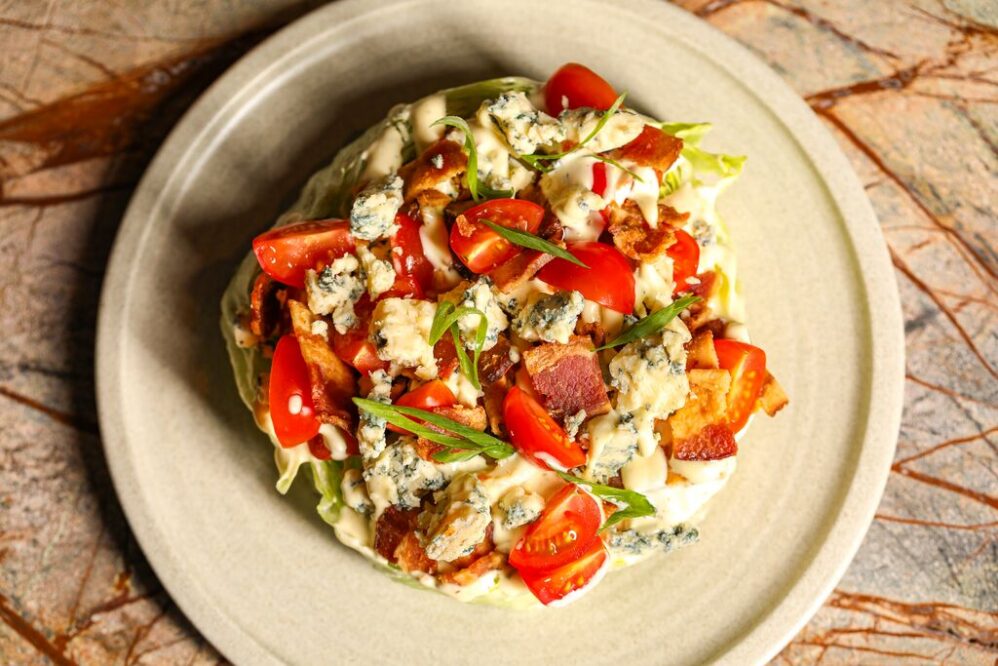 A wedge salad topped with cherry tomatoes, blue cheese crumbles, crispy bacon, chopped green onions, and creamy dressing, served on a beige plate over a marbled surface.