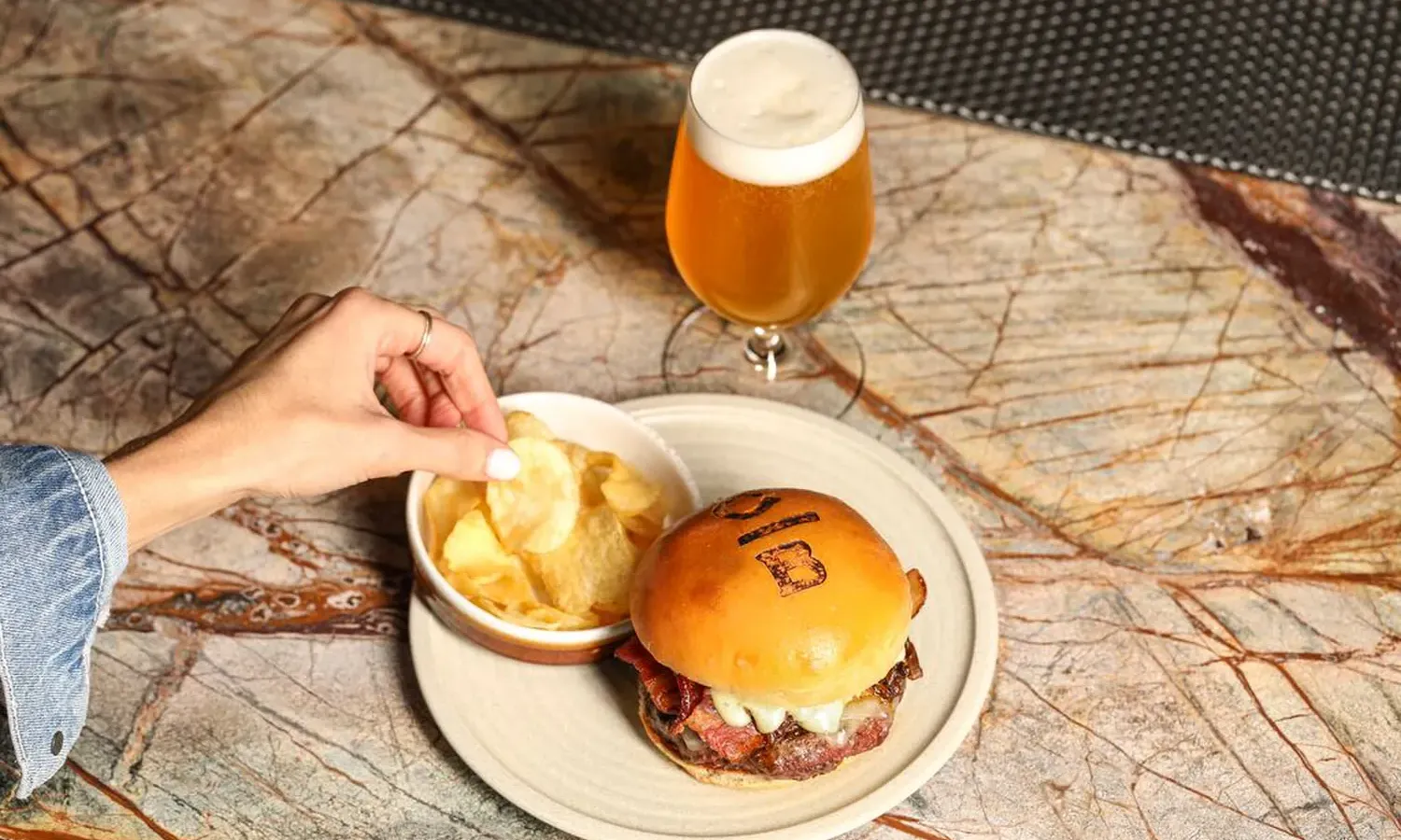 A hand reaches for potato chips in a bowl next to a burger with a branded bun on a plate. A glass of frothy beer sits behind the plate on a marbled table surface.