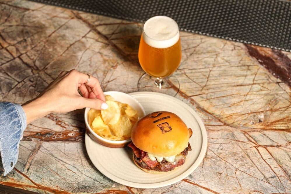 A hand reaches for potato chips in a bowl next to a burger with a branded bun on a plate. A glass of frothy beer sits behind the plate on a marbled table surface.