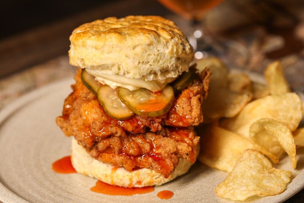 A fried chicken biscuit sandwich with pickles and sauce, topped with mayonnaise, served on a plate with potato chips on the side.
