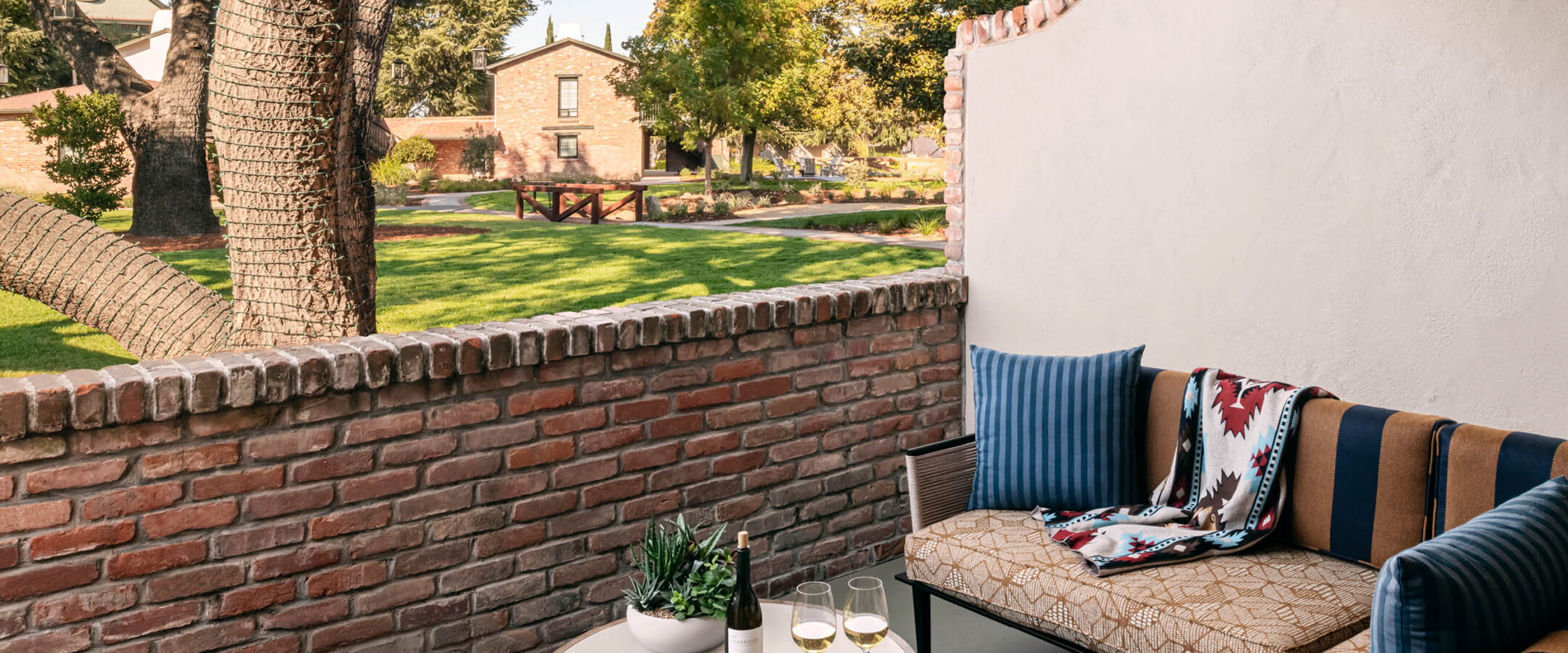 A cozy patio with a cushioned bench, striped and patterned pillows, a blanket, a small table holding wine, glasses, and sunglasses, overlooks a grassy courtyard with trees and a brick building in the background.
