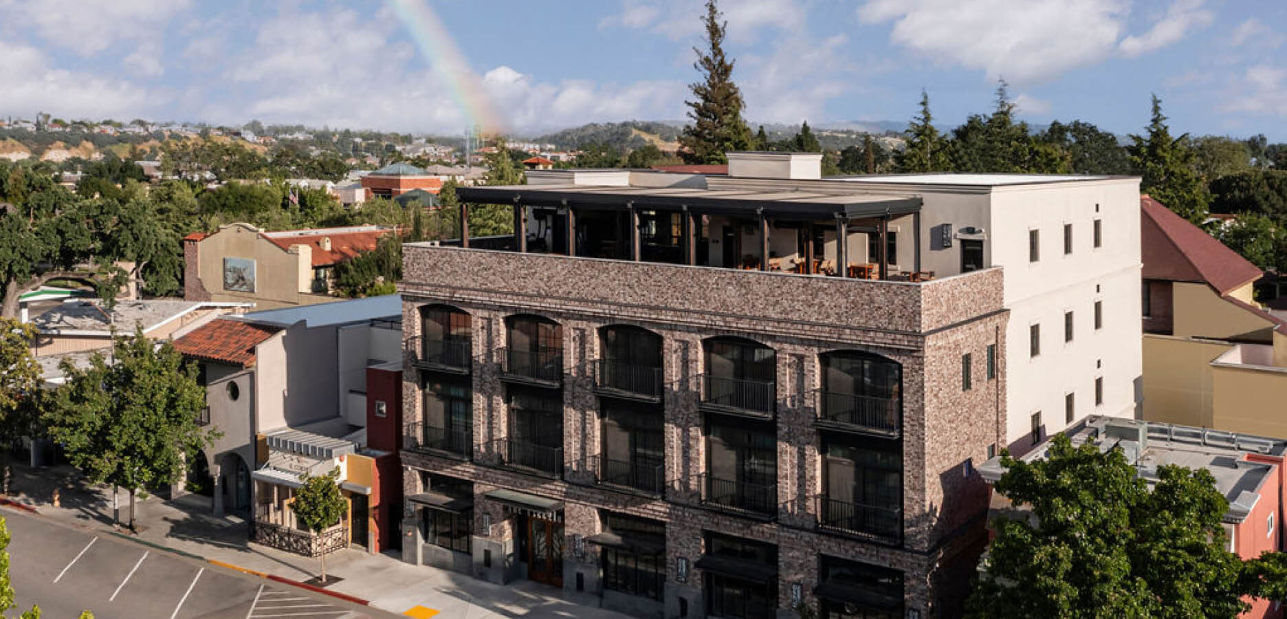 A three-story brick building with large windows and a rooftop patio sits on a quiet street lined with trees. In the background, a faint rainbow arches over a suburban neighborhood under a partly cloudy sky.