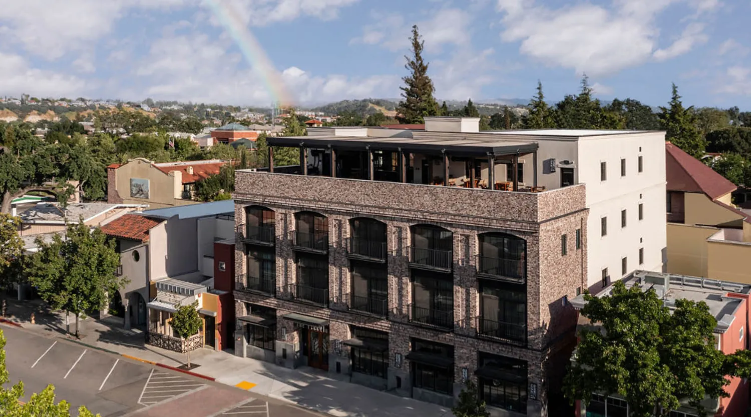 A three-story brick building with large windows and a rooftop patio sits on a quiet street lined with trees. In the background, a faint rainbow arches over a suburban neighborhood under a partly cloudy sky.