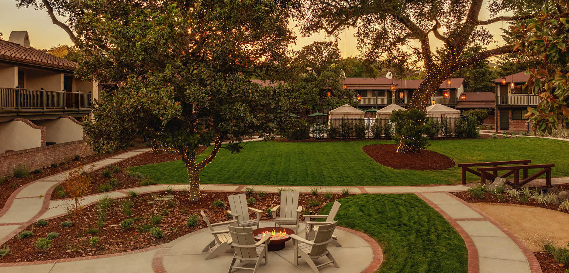 A landscaped courtyard at sunset with green lawns, mature trees, a fire pit surrounded by Adirondack chairs, walking paths, and two-story buildings with balconies in the background.
