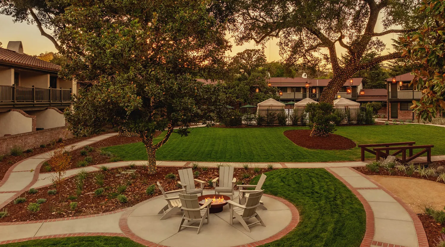 A landscaped courtyard at sunset with green lawns, mature trees, a fire pit surrounded by Adirondack chairs, walking paths, and two-story buildings with balconies in the background.