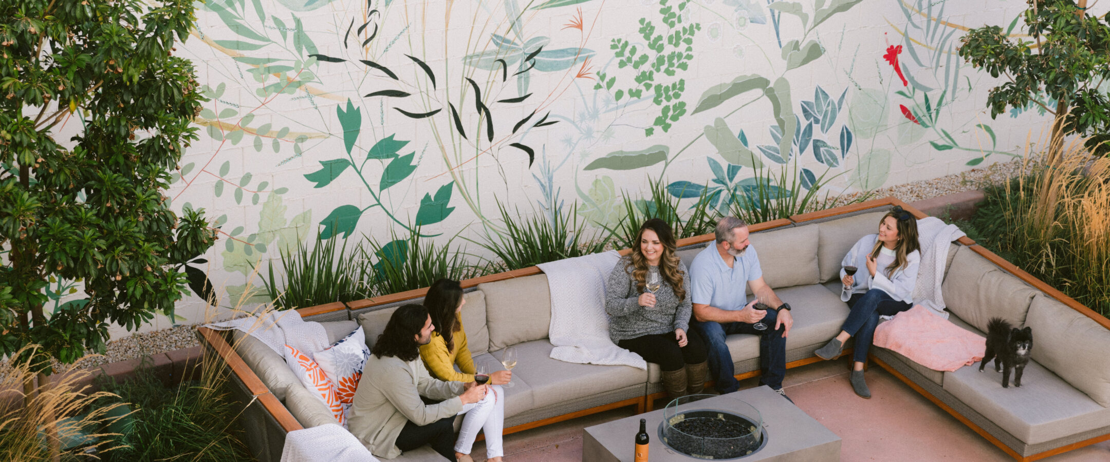 Four people sit on outdoor couches around a fire pit, chatting and holding drinks, with a dog nearby. Behind them is a large mural featuring green plants and leaves. A wine bottle sits on the table.