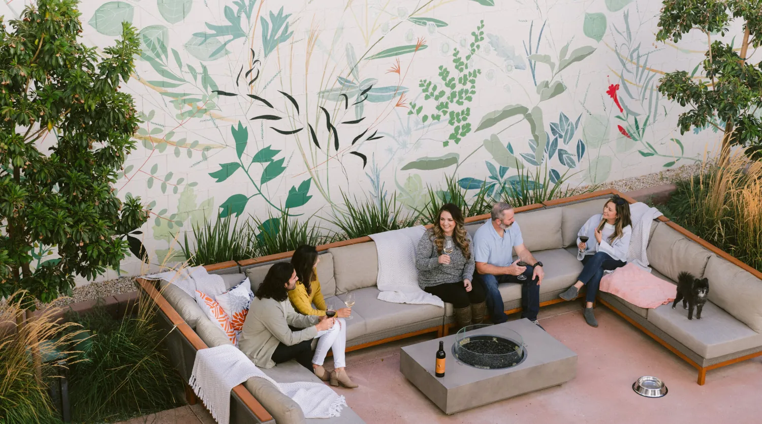 Four people sit on outdoor couches around a fire pit, chatting and holding drinks, with a dog nearby. Behind them is a large mural featuring green plants and leaves. A wine bottle sits on the table.