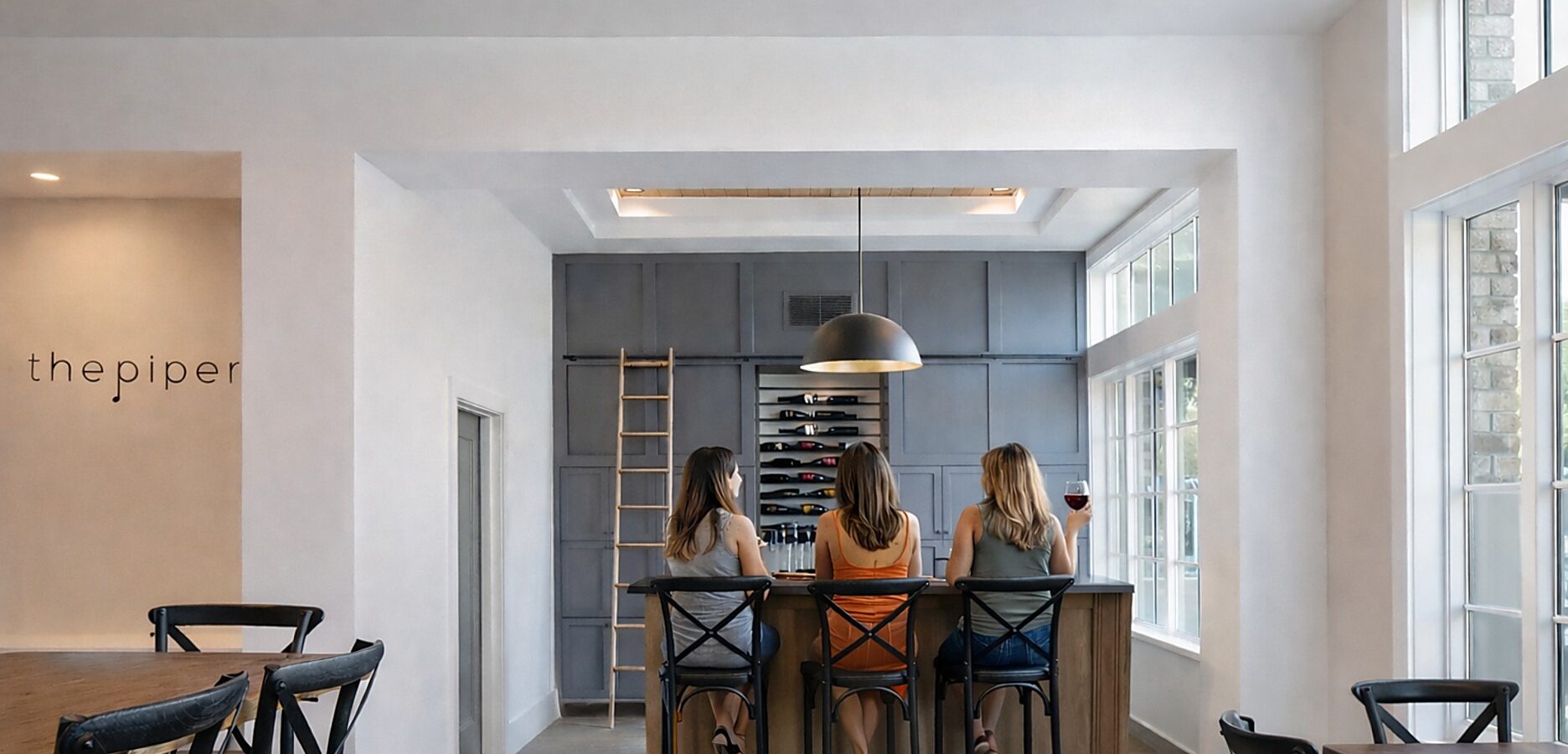 Three women sit at a bar in a modern, bright room at The Piccolo at Paso Robles Inn, with large windows, a wine rack on the back wall, and a ladder leaning against it. They each have a drink and are engaged in conversation.