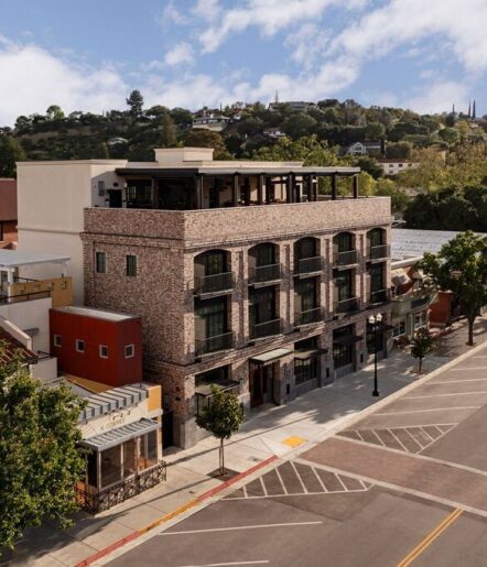 A three-story brick building with a rooftop terrace stands on a quiet street lined with trees and colorful smaller buildings, with a hillside neighborhood and blue sky in the background.