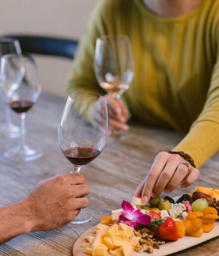 Two people sit at a table with three glasses of wine and a wooden platter of cheese, nuts, fruits, and flowers. One person reaches for food while the other holds a glass of red wine.