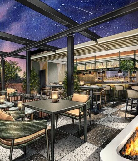Rooftop bar and lounge with modern furniture, tables, and barstools, softly lit by lanterns. The space features an open ceiling, showing a starry night sky, and a cozy fire pit in the foreground.