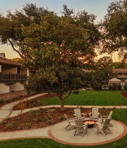 Four white Adirondack chairs surround a fire pit on a circular patio, set in a landscaped backyard with green grass, planted beds, a large tree, and winding pathways at sunset.