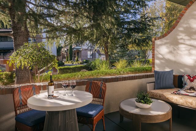 A cozy outdoor patio with a round table set for two with wine, wicker chairs, a cushioned bench with pillows, potted plants, and a view of trees and buildings in the background.