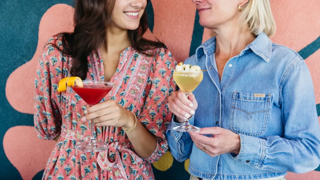 Two women smiling and holding cocktails stand in front of a colorful floral mural at Salina Rooftop. One wears a pink patterned dress with a red drink, while the other sports a denim shirt and holds a yellow cocktail.