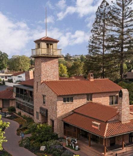 A brick building with a tall tower and red-tiled roofs sits among trees; signs read MacArthur and Park. A cow statue is near the entrance, and a blue sky with clouds is visible overhead.