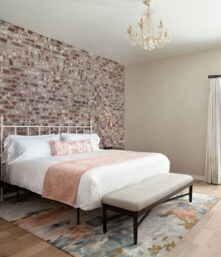 A modern bedroom with an exposed brick accent wall, white metal bed frame, white bedding with a pink throw, a bench at the foot of the bed, colorful rug, chandelier, and cream-colored walls with curtains.