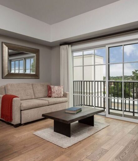 A modern living room with a beige sofa, red throw, large mirror, dark coffee table, light rug, and floor-to-ceiling windows overlooking a balcony with a scenic view. The room has wood flooring and neutral tones.