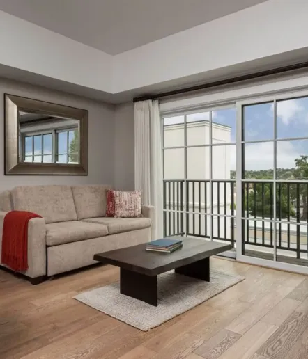 A modern living room with a beige sofa, red throw, large mirror, dark coffee table, light rug, and floor-to-ceiling windows overlooking a balcony with a scenic view. The room has wood flooring and neutral tones.