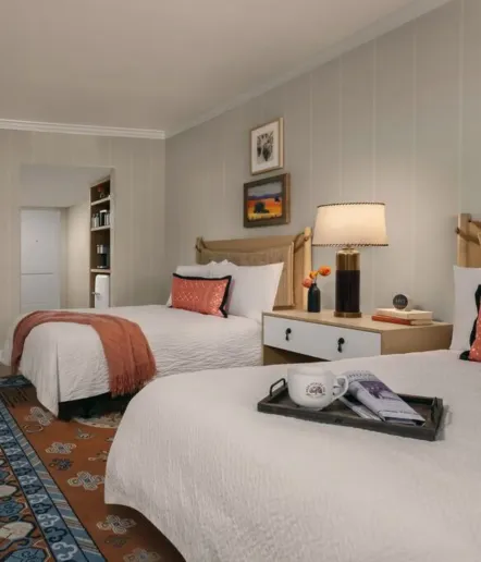 A cozy hotel room with two beds, white bedding, a nightstand with a lamp, flowers, and books. A tray with a coffee cup and magazine sits on one bed. Art hangs on the pale wall, and a colorful rug lies on the floor.