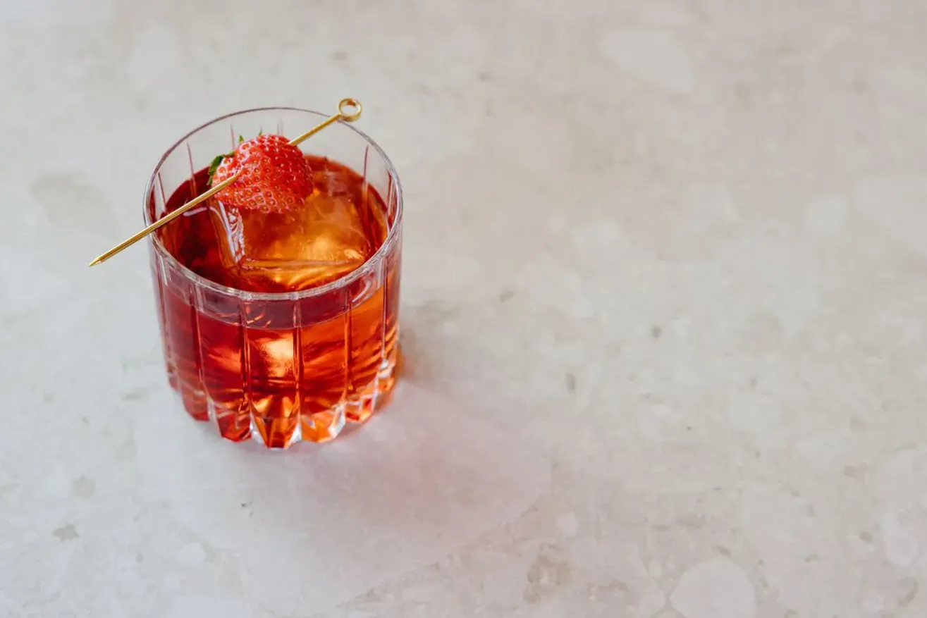 A glass of red cocktail with ice sits on a light-colored stone surface, garnished with a strawberry on a cocktail pick.