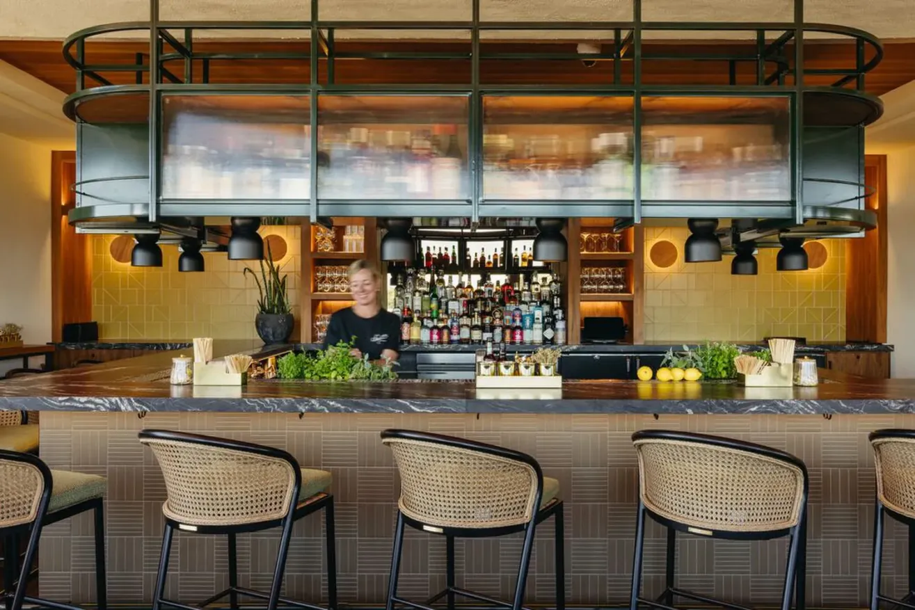 A bartender stands behind a modern bar with a marble countertop, greenery, and bottles of liquor on shelves. Four woven-back barstools are at the counter, and warm lighting creates a welcoming atmosphere.