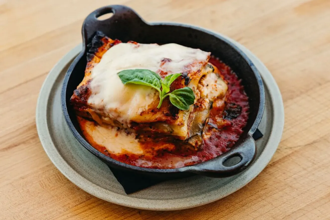 A slice of lasagna topped with melted cheese and fresh basil sits in a small cast iron skillet on a plate, with rich tomato sauce pooling around the edges.